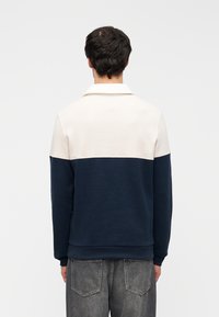 Φούτερ μπλουζάκι σε μπλε ναυτικό και κρεμ χρώματα με γιακά, με μαλακή υφή και άνετη εφαρμογή. Η πίσω όψη δείχνει αντιθετικά χρώματα.