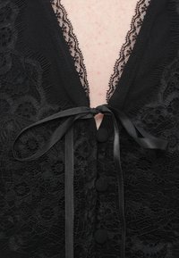 Μαύρο κορμάκι από δαντέλα με βαθύ V λαιμόκοψη, σατέν κορδέλα για δεσίματα και διακοσμητικά κουμπιά στην μπροστινή πλευρά. Μαλακό, υφασμάτινο με περίτεχνα σχέδια.