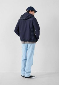 Marineblauwe jas met een gewatteerde capuchon en zijkanten, gecombineerd met lichtblauwe jeans en zwarte sneakers, met een casual, ontspannen pasvorm.