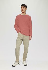 Rosa Sweatshirt mit langen Ärmeln und rundem Ausschnitt, kombiniert mit hellbeigen Jeans und neutralen Sneakers mit grauen und grünen Akzenten.