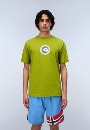 Giovane uomo con capelli ricci che indossa una maglietta verde lime e pantaloncini blu con un motivo a bandiera rossa, bianca e blu, in piedi contro uno sfondo chiaro.