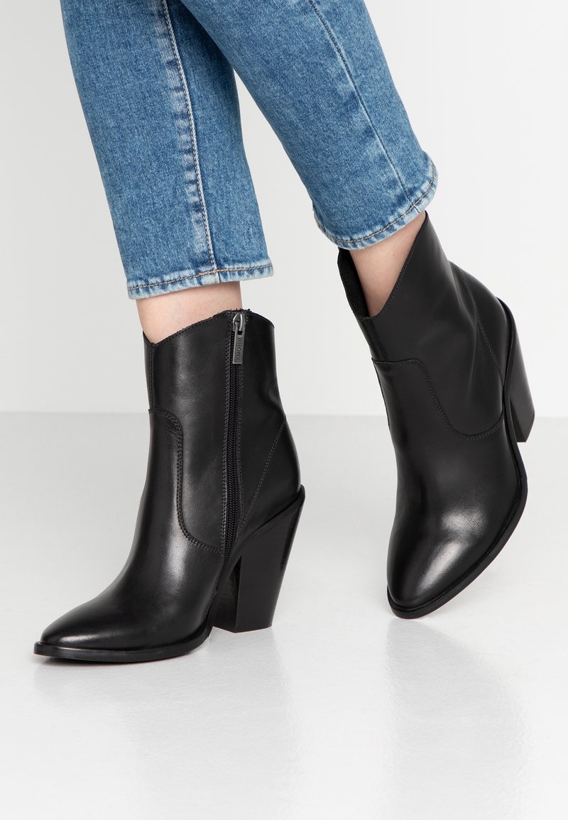 Botines de cuero negro hasta el tobillo con punta afilada, cierre de cremallera y un tacón de bloque alto, combinados con jeans de mezclilla azul con vuelta.