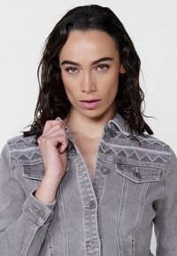 Siva denim jakna s kompleksno teksturno vezenje na ramenih, z gumbi in žepi na prsih. Gladka površina, rahlo prilegajoča.