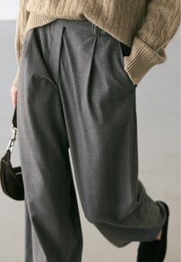 Pantalon gris plissé à fines rayures verticales, en tissu texturé. Doté d'un passant pour ceinture et d'une coupe décontractée, associé à un pull en tricot beige.