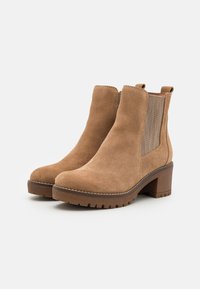Zamszowe ankle boots w kolorze tan, z elastycznymi bocznymi panelami, uchwytami do zakładania, grubymi, niskimi obcasami i teksturowanymi gumowymi podeszwami na białym tle.
