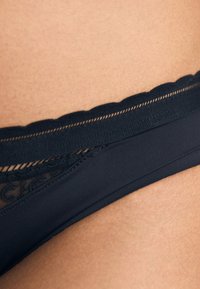 Črne bikini hlačke z prozornimi čipkastimi detajli, valovitim elastikastim pasom, gladkim materialom in brezšivnimi robovi. Poudarek na oblikovalskih podrobnostih.