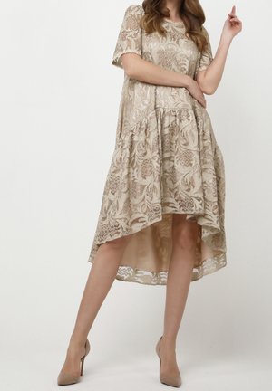 Vrouw draagt een beige kanten jurk met een hoge-lage zoom, korte mouwen en nude hoge hakken, poserend met een gebogen arm en opgestoken hand.