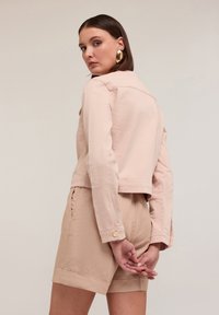 Ljusrosa kort jacka med krage, axelsömmar och knappade manschetter, i kombination med beige shorts som har en vikning vid fållen.