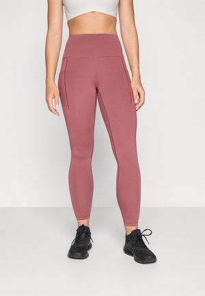 Leggings taille haute et ajustés en tissu rose poussiéreux, avec deux poches latérales et des coutures renforcées, associés à des chaussures de sport noires.