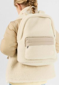 Kind met gevlochten haar, gekleed in een beige fleecejack en een bijpassende, pluizige crème rugzak, kijkt weg tegen een effen achtergrond.