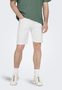 Bermudas de mezclilla blancas con puños enrollados, combinadas con una camiseta verde holgada y zapatillas blancas. Tejido texturizado y ajuste casual.