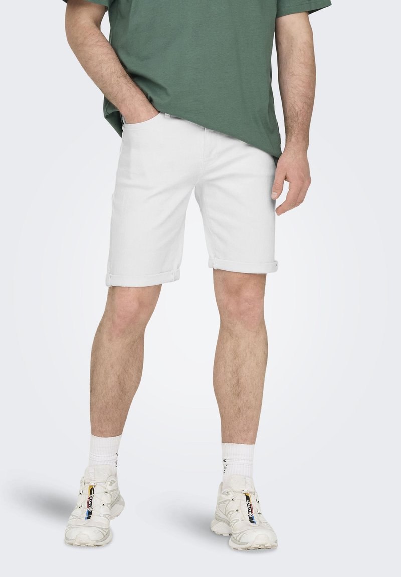 Bermudas de mezclilla blancas con puños enrollados, combinadas con una camiseta verde holgada y zapatillas blancas. Tejido texturizado y ajuste casual.