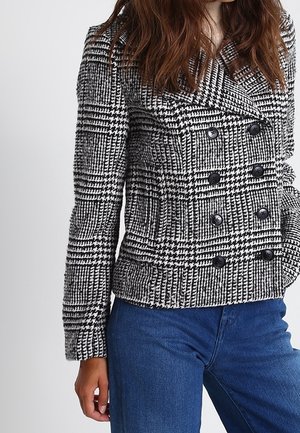 Kobieta ubrana w czarno-biały żakiet dwurzędowy w jodełkę z dużymi guzikami i niebieskie dżinsy, stojąca z jedną ręką w kieszeni.