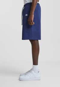 Navy blue shorts with an elastic waistband and drawstring, featuring a logo patch on the side. Paired with white sneakers and white socks.