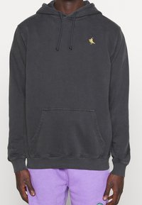 Sudadera con capucha de algodón gris oscuro con un bolsillo frontal, capucha con cordón y un pequeño emblema de pájaro amarillo en el pecho. Textura suave.