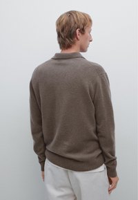 Brown knitted pullover sweater with a ribbed collar, cuffs, and hem, featuring a relaxed fit and soft texture. Worn over cream-colored trousers.