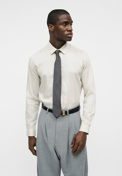 Άνδρας με λευκό πουκάμισο με λεπτές κάθετες ρίγες, γκρι γραβάτα, γκρι πλισέ παντελόνι και μαύρη ζώνη, που κοιτάζει προς τα δεξιά του.
