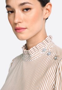Bluzka w paski z falbanianym kołnierzem w kolorze beżowym i białym. Z ozdobnymi guzikami z perłowymi akcentami na lewym ramieniu. Gładka faktura.
