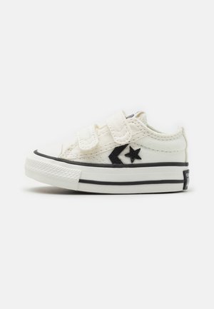 White canvas shoes with black accents, featuring Velcro straps, a rubber sole, and a star logo on the side. Rounded toe design.