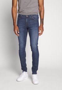 Jeans ajustados de mezclilla azul oscura con un ligero desgastado, que cuentan con un diseño de cinco bolsillos, cierre de cremallera y trabillas para el cinturón, combinados con zapatillas blancas.