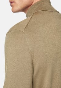 Beige knit turtleneck sweater with a close-fitting design, ribbed texture, and a high collar. Features soft fabric and shoulder seams.