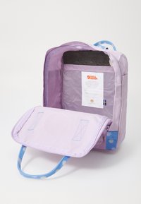Mochila roxa com uma abertura com fecho, tecido macio, compartimento interior com um painel cinza, alças azuis com um padrão de bolinhas e um rótulo.
