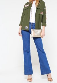 Olivenfarvet knap-til-jakke med lapper, hvid skjorte, blå flared jeans, guldfarvet clutch med frynse, nude sandaler.