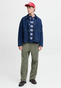 Denimjakke med knappelukking, stripete strikket skjorte under, olivengrønne kordfløyelsbukser og rød lue. Brune sko fullfører antrekket.