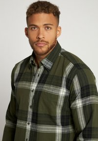 Camicia di flanella a quadri verdi e neri con bottone frontale, maniche lunghe e tasca applicata. Presenta un tessuto testurizzato e un classico design del colletto.