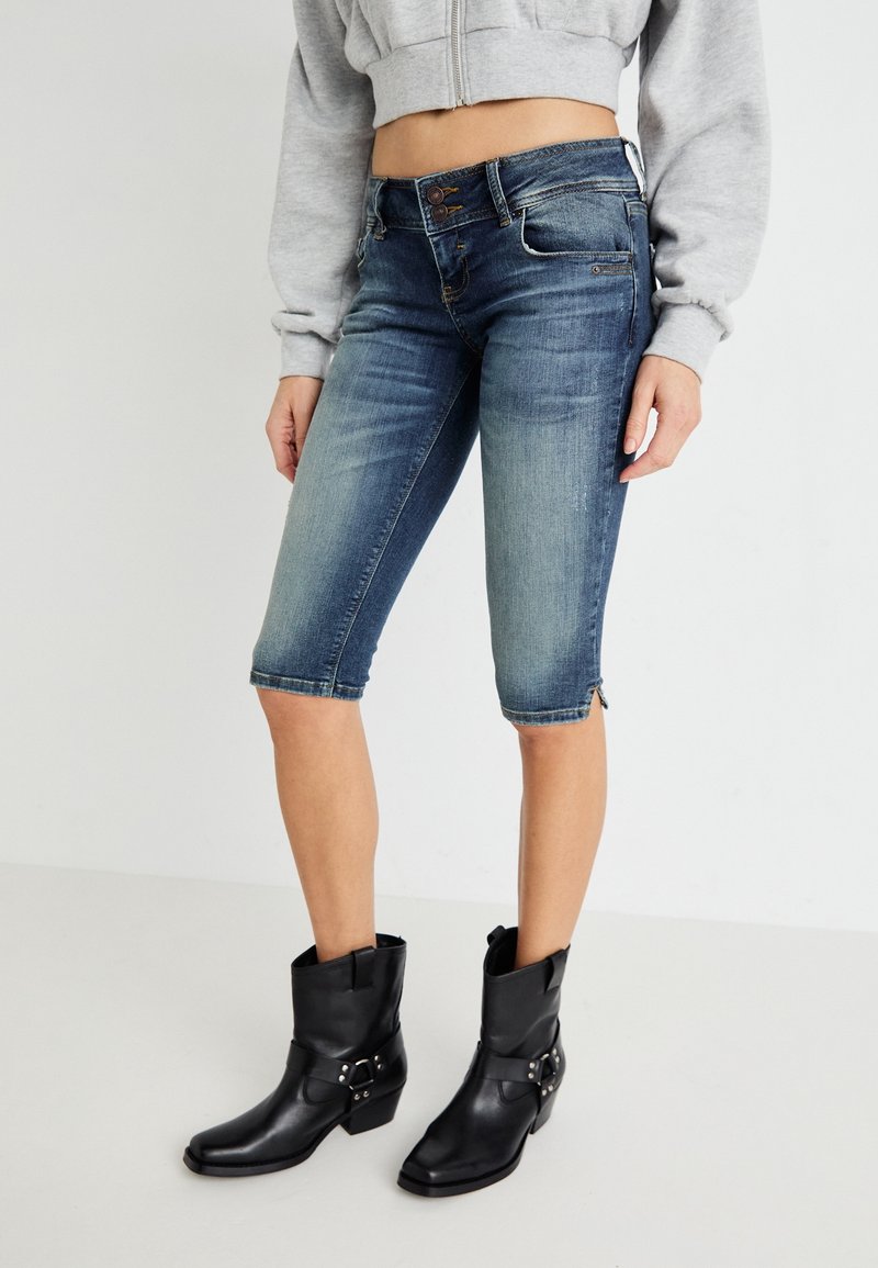 Femme portant un short en jean bleu mi-long, un sweat-shirt gris court et des bottines noires style cowboy, debout devant un fond blanc.