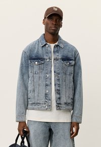 Φωτεινό γαλάζιο τζιν μπουφάν με κούμπωμα κουμπιών, δύο τσέπες στο στήθος και διακριτική ξεθώριαση. Φορεμένο πάνω από λευκό μπλουζάκι, σε συνδυασμό με τζιν παντελόνι.