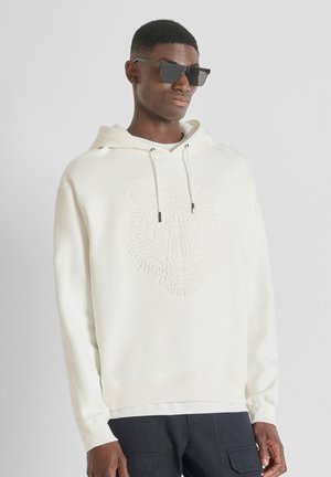 Cremefarbener Kapuzenpullover mit einem strukturierten Löwenkopfmuster in der Mitte, mit Kordelzugkapuze und gerippten Bündchen und Saum. Getragen mit dunkler Sonnenbrille.