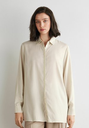 Frau mit schulterlangem dunklem Haar, die ein cremefarbenes Hemd mit langen Ärmeln trägt und vor einer weißen Wand steht.