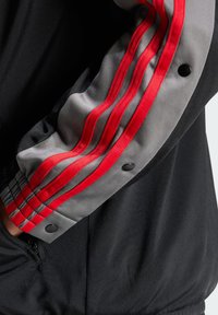 Veste de sport noire avec des accents rayés gris et rouge sur les manches, dotée d'une finition texturée et de boutons-pression noirs.