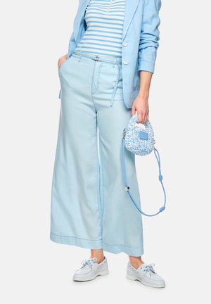 Frau trägt hellblaue, weit geschnittene Hosen, passenden Blazer, gestreiftes Oberteil, weiße Bootsschuhe und hält eine kleine blau-weiße, gewebte Handtasche.