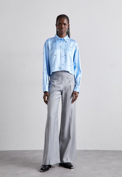 Cămașă scurtă de culoare bleu deschis, cu accesorii decorative din argint, asociată cu pantaloni gri cu talie înaltă și picioare largi, și încălțăminte neagră.