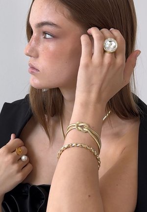 Guldsmycken med en stor klar stenring, sammanflätade armband och en pärlring. Släta texturer med varierade ytor. Svart topp.