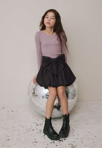 Top viola a maniche lunghe abbinato a una gonna nera con un grande fiocco in vita. Stivaletti neri completano l'outfit.