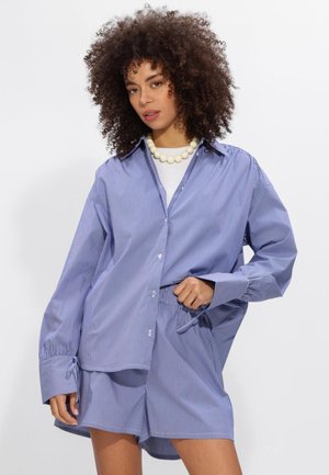 Vrouw met krullend haar, gekleed in een oversized shirt met blauwe en witte strepen en bijpassende korte broek, afgewerkt met een witte kralenketting.