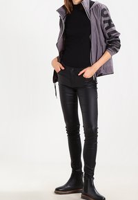 Žena oblečená v šedé zipové bundě, černém dlouhém rukávu, černých kožených kalhotách a černých kotníkových botách, stojí s rukama v kapsách.