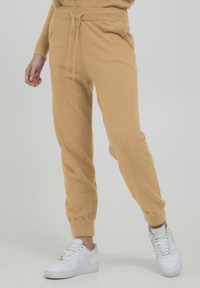 Tan joggerbukser lavet af blødt stof, med snøre i taljen, tætsiddende manchetter og sidelommer, parret med hvide sneakers.