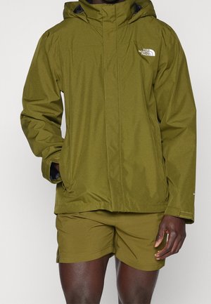 Person trägt eine olivgrüne, wasserdichte The North Face Jacke mit angesetzter Kapuze und passende Shorts, steht vor schlichtem Hintergrund.