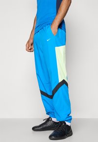 Pantalones deportivos azules con un acento de malla negra y verde, que presentan una cinturilla elástica y puños ajustados. Combinados con zapatillas deportivas negras.