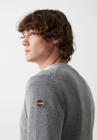 Maglione grigio lavorato a maglia con un patch rotondo sul lato sinistro della spalla. Presenta un collo rotondo e un tessuto testurizzato per il calore.