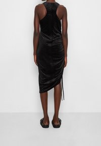 Osoba stojí zády k divákovi, má na sobě černé bezrukávové šaty se strukturovaným vzorem a nazouvací sandály.