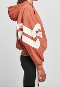 Orange lättviktsjacka med huood och vita chevronränder, elastiska ärmar och avslappnad passform. Slät yta och enkel design.