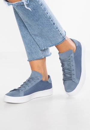 Jambes croisées portant un jean bleu clair à ourlet effiloché et des baskets bleues à lacets avec semelles blanches sur une surface blanche.