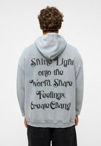 Grijs, hoodie sweatshirt met zwarte tekst op de achterkant: "Laat Licht op de Wereld Schijnen. Deel Gevoelens, Creëer Verandering." Zachte stof, relaxte pasvorm.