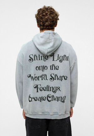 Grijs, hoodie sweatshirt met zwarte tekst op de achterkant: "Laat Licht op de Wereld Schijnen. Deel Gevoelens, Creëer Verandering." Zachte stof, relaxte pasvorm.