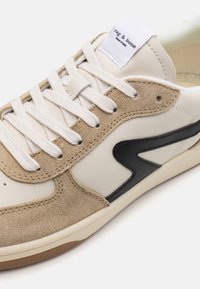 Beżowe skórzane i zamszowe sneakersy z białymi sznurówkami, czarnym akcentem i logo na języku. Gumowa podeszwa o lekkiej teksturze.